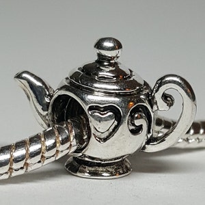 Könnte beinhalten: Ein silberner Charm in Form einer Teekanne. Der Charm hat einen kleinen Griff, einen Ausguss und einen Deckel. Der Körper der Teekanne hat herzförmige Ausschnitte. Der Charm ist an einem silberfarbenen Armband befestigt.