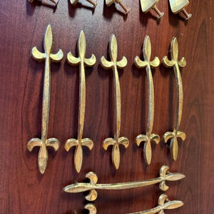 May include: A collection of vintage brass hardware, including six fleur-de-lis handles, six triangular knobs, and two smaller handles. The hardware is arranged on a dark wood surface, showcasing its aged, golden finish.