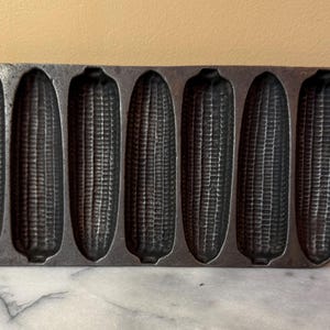 May include: A cast iron corn stick pan with six corn-shaped molds. The pan is dark gray and has a handle on each end. The molds are detailed to resemble ears of corn, perfect for baking corn bread.