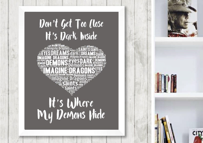 May include: A gray and white printable wall art design with a heart-shaped word cloud. The text in the word cloud includes "Imagine Dragons", "Demons", "Saints", and "Dreams". The text below the word cloud reads "Don't Get Too Close It's Dark Inside It's Where My Demons Hide".