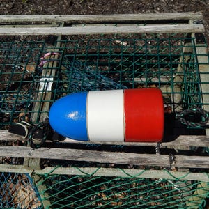 May include: A blue, white, and red buoy rests on a wooden lobster trap. The buoy is cylindrical with a rounded top and is painted with a blue top, white center, and red bottom. A black rope is attached to the buoy.