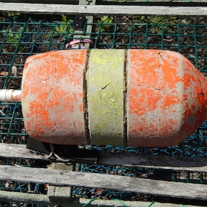 May include: A cylindrical marine buoy featuring a weathered, cracked orange finish on the ends and a central band of faded yellow. The buoy is constructed from three distinct segments and includes a white plastic rod extending through the center.