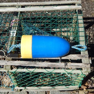 May include: A blue and yellow buoy with rope attached, resting on a wooden lobster trap. The buoy is cylindrical with a rounded top and a yellow top. The trap is made of wood and green netting.