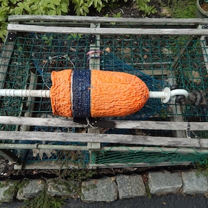 May include: A rectangular wooden lobster trap with a green mesh netting interior, featuring a textured orange and dark blue cylindrical buoy attached to a white plastic rod and a black rope, resting horizontally across the top of the wooden frame.