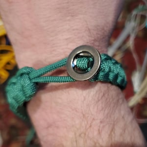 May include: A green paracord bracelet with a silver metal clasp. The bracelet is tied with a knot and has a simple design.