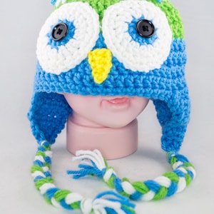 May include: A blue, green, and white crocheted owl hat with a long braided strap. The hat has large white eyes with black buttons and a yellow beak.