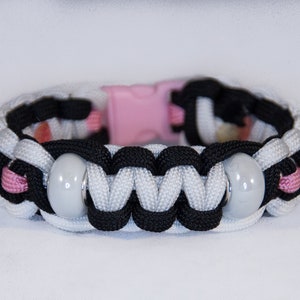 May include: A handmade paracord bracelet featuring a woven design in black, white, and pink. The bracelet has two gray beads and a pink plastic clasp. This accessory is suitable for everyday wear.