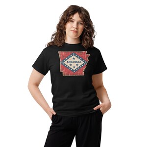 Puede incluir: Una camiseta negra de manga corta con un gráfico del estado de Arkansas en un patrón de bandera desgastado de color rojo, blanco y azul. El diseño incluye la palabra ARKANSAS en letras azules centrada dentro de un rombo blanco.