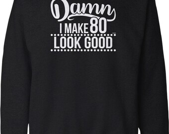 May include: A black long-sleeve crewneck sweatshirt featuring white screen-printed text that reads Damn I MAKE 80 LOOK GOOD, with the word Damn in a script font and the remaining text in a bold sans-serif typeface.