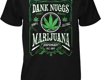 May include: A black short-sleeve t-shirt featuring a green and white graphic print. The design includes the text DANK NUGGS, FINEST QUALITY, 100% NATURAL, MARIJUANA, DISPENSARY, and EST. 1997, centered around a large cannabis leaf illustration.