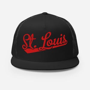 May include: A black trucker hat featuring a mesh back and a flat brim. The front panel displays the text St. Louis embroidered in a bold red script font with a red underline accent. The cap has a structured crown and a solid black front panel.
