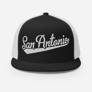 May include: A black and white trucker hat featuring a flat brim and a mesh back. The front panel is black with the text San Antonio embroidered in white cursive script, underlined by a white swoosh design across the center.