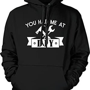 May include: A black hooded sweatshirt featuring a white graphic print on the chest. The design includes a crossed hammer and wrench above a banner containing the text "YOU HAD ME AT DIY" in a bold, white, sans-serif font with a front pocket.