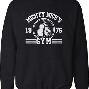 May include: A black long-sleeve sweatshirt featuring a white graphic print on the chest. The design includes two boxing gloves inside a circle, with the text MIGHTY MICK'S, 1976, and GYM arranged around the central emblem.