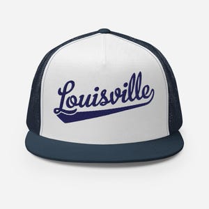 May include: A white and navy blue trucker hat featuring a structured front panel with the word Louisville embroidered in navy blue script font. The cap includes a solid navy blue flat brim and navy blue mesh side and back panels.