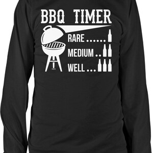 May include: Black long-sleeve shirt with a white graphic. The graphic includes the text "BBQ TIMER" above a grill illustration and the words "RARE," "MEDIUM," and "WELL" with bottle icons. A fun shirt for grilling enthusiasts.
