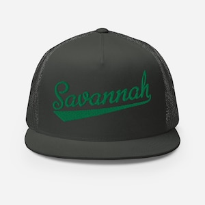 May include: A charcoal gray trucker hat featuring a structured front panel and a mesh back. The word Savannah is embroidered in green cursive script across the front, underlined by a solid green swoosh accenting the design.