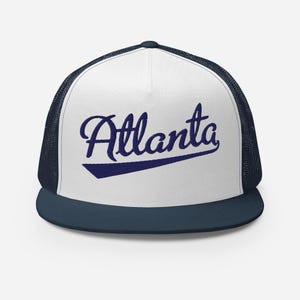 May include: A white and navy blue trucker hat featuring a structured front panel with the word Atlanta embroidered in a dark blue cursive script font, underlined by a solid navy blue swoosh, and a breathable mesh back with a flat brim.