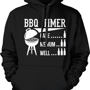 May include: Black hooded sweatshirt with white graphic text and illustrations. The text reads "BBQ TIME" above a grill illustration, followed by "RARE... MEDIUM... WELL..." with bottle illustrations. A casual, comfortable garment.