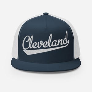 Könnte beinhalten: Eine marineblaue und weiße Trucker-Cap mit einem strukturierten Vorderteil, auf dem das Wort Cleveland in weißer Schreibschrift gestickt ist. Das Design umfasst einen flachen Schirm und ein atmungsaktives Netzrückenteil für einen klassischen Look.