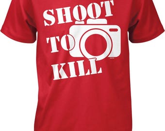 May include: A red short-sleeved t-shirt featuring a white graphic of a camera and the text "SHOOT TO KILL" printed in a bold, white, sans-serif font. The camera icon is positioned centrally, with the text arranged around it.