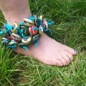 May include: A blue and green ankle bracelet made from bottle caps. The bracelet is worn on a person's ankle, and the person's foot is resting on green grass.