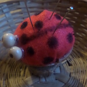 Op de afbeelding: Een rood en zwart lieveheersbeestje-vormig naaldenkussen met witte knuffelogen en verschillende spelden die eruit steken. Het naaldenkussen ligt in een geweven mand.