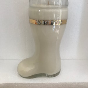 May include: A white candle shaped like a beer boot with a gold band around the top that says "Warsteiner".