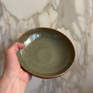 May include: A handmade ceramic bowl with a speckled green interior and a brown rim. The bowl is held in a hand, showcasing its shape and texture. The bowl is likely used for serving food.