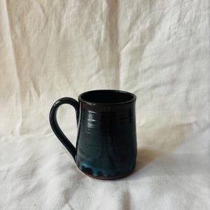 May include: A ceramic mug with a glossy finish, featuring a deep black glaze that transitions into a vibrant teal blue drip pattern near the base. The mug has a curved handle on the left side and a smooth, cylindrical body with a visible terracotta rim.