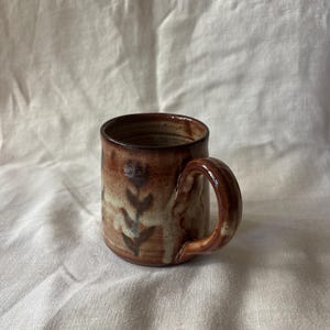 May include: A cylindrical ceramic mug with a glossy, mottled brown and cream glaze. The exterior features a dark brown, hand-painted botanical motif of a vertical stem with leaves. It has a sturdy, curved handle and a smooth, rounded rim.