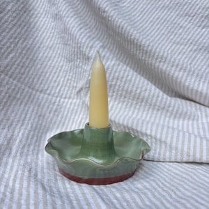 May include: A handmade ceramic candle holder with a green top and a red base, holding a tall, slender, cream-colored candle. The candle holder has a ruffled edge and sits on a striped, neutral-toned fabric.