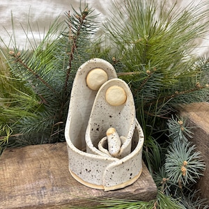 May include: A ceramic nativity scene with a speckled, off-white finish. The set includes figures of Mary, Joseph, and baby Jesus, each with a circular accent. The scene is set on a wooden surface, surrounded by evergreen branches.