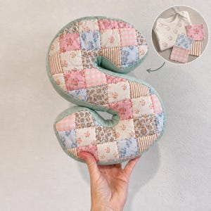 May include: A plush letter S pillow constructed from a patchwork of fabric squares featuring floral, gingham, and striped patterns in shades of pink, blue, and cream. A circular inset shows a matching baby bodysuit and three small fabric squares.