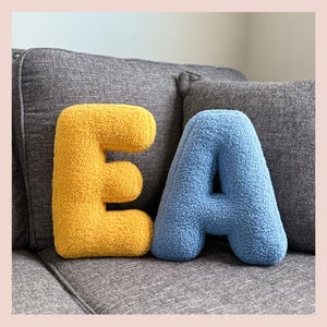 May include: Two plush, textured letter pillows, a yellow "E" and a blue "A", rest on a gray sofa. The letters are soft and fluffy, adding a decorative touch. The background includes gray cushions and a light pink border.