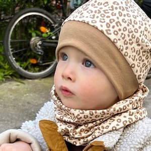 Könnte beinhalten: Ein Baby mit einer braunen und beigen Leopardenmuster-Mütze und einem passenden Halstuch. Die Mütze hat einen einfarbigen beigen Rand. Das Baby trägt eine weiße und beige Jacke.