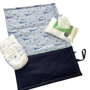 Può includere: Un fasciatoio blu navy e fantasia con un design a balene e creature marine. Include un pannolino e un pacchetto di salviette. Il tappetino è aperto, rivelando il motivo interno.