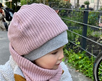 May include: A child wearing a pink knitted hat and matching scarf. The hat has a grey brim and the scarf is folded around the neck.