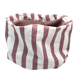 May include: A circular neck warmer featuring a vertical striped pattern in muted mauve and off-white. The interior lining is a solid dark brown fabric. The text "kinnerleev" with a small heart icon is printed in black in the bottom right corner.