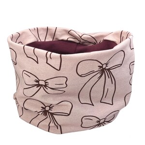 May include: A light pink neck warmer featuring a repeating pattern of brown bows. The inner lining is a deep burgundy colour. The neck warmer is made of soft fabric and is designed to be worn around the neck.