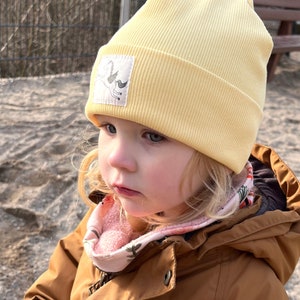 May include: A young child wearing a yellow beanie with a unicorn patch and a brown jacket with a pink and floral patterned scarf.