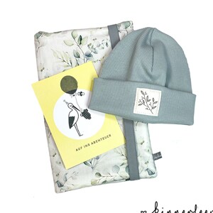 May include: A baby gift set. It includes a light blue ribbed knit beanie with a small square patch and a botanical design. The set also features a blanket with a botanical print and a yellow card with a stork illustration.
