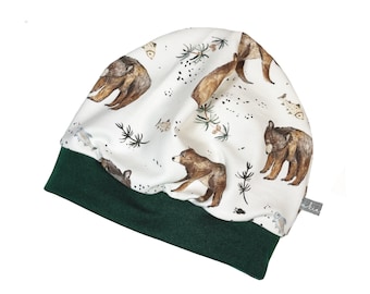 Puede incluir: Un gorro blanco con un borde verde presenta un patrón repetido de osos marrones y pinos. El gorro tiene una pequeña etiqueta con la palabra "kinnerlee" en él.