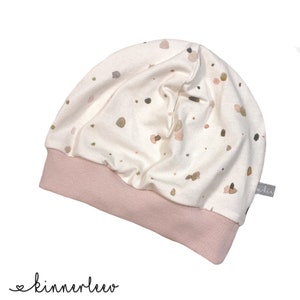 May include: A white beanie with a pink fold-over brim and a scattered watercolour print of brown, green, and pink dots. The beanie has a small label with the word "skin" on it.