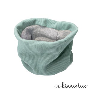 Könnte beinhalten: Ein salbeigrüner, gerippter Schlauchschal mit einem weichen, grau melierten Innenfutter. Das Accessoire hat eine kreisförmige, röhrenförmige Konstruktion mit einer strukturierten Strickaußenseite und einem glatten Innenfutter.