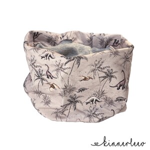 Könnte beinhalten: Ein beiger Halsschmeichler mit einem Muster aus Palmen und Dinosauriern. Der Halsschmeichler ist aus einem weichen, dehnbaren Stoff gefertigt.