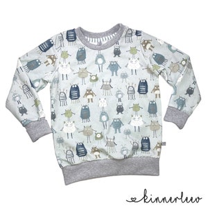 Peut inclure: Un sweat à manches longues bleu clair avec une bordure grise. Le sweat-shirt présente un motif répété de monstres de dessin animé colorés.