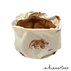 Könnte beinhalten: Ein beigefarbener Stoffaufbewahrungskorb mit brauner Auskleidung. Der Korb hat ein sich wiederholendes Muster aus braunen Bären und Fischen auf weißem Hintergrund.