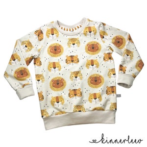 Può includere: Una felpa a maniche lunghe color crema con un motivo ripetuto di volti di leone arancioni e marroni. La felpa ha un collo a girocollo e polsini a costine.