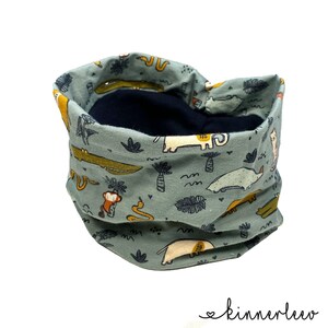 Könnte beinhalten: Ein blauer und grüner Stoff-Halswärmer mit einem Muster aus Tieren und Pflanzen. Der Halswärmer ist reversibel und hat ein schwarzes Fleece-Futter.